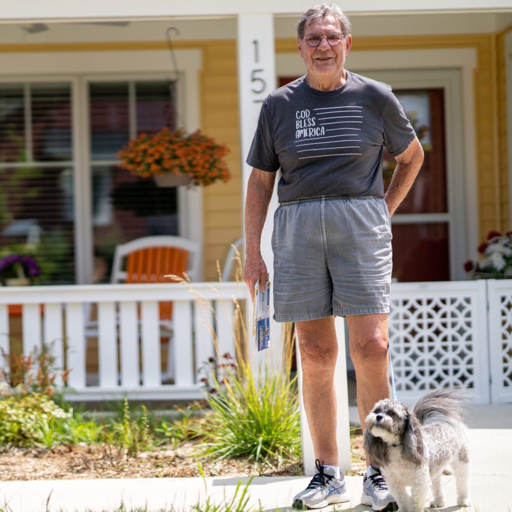 DSC06523 An older gentleman stands in front of the front porch of a charming yellow cottage. He looks warmly at the camera and is holding mail, and he wears a gray shirt with the text "God bless America." In front of him, a small gray-and-white dog on a leash.