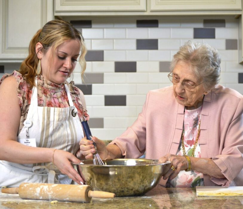 Screenshot A younger woman in a white apron holds a stainless steel bowl while an older woman in a pink jacket stirs with a whisk. They are surrounded by baking supplies.