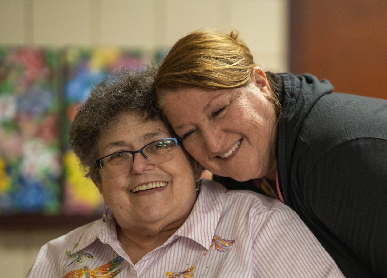 Screenshot Two women, one sitting and one standing, embrace closely while smiling at the camera. The woman sitting is slightly older and wears a pink striped dragonfly shirt and glasses. The woman standing is a bit younger and has brown hair and freckles.